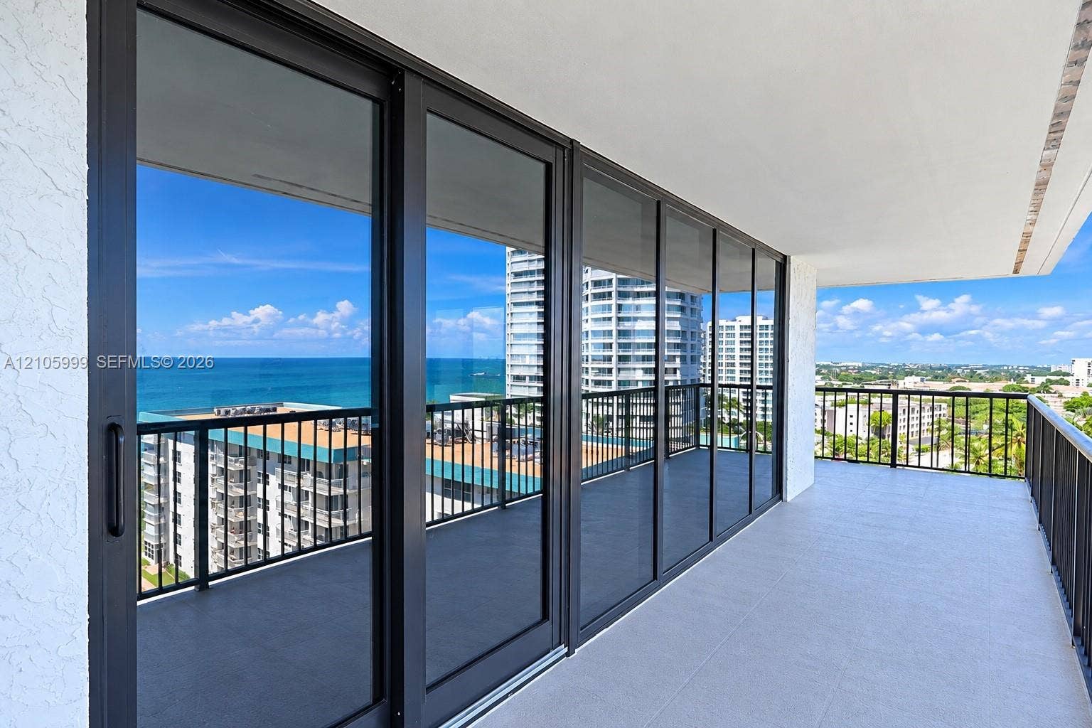 Spacious wrap-around balcony featuring large-format tile flooring, black metal railings, and floor-to-ceiling sliding glass doors with dark frames