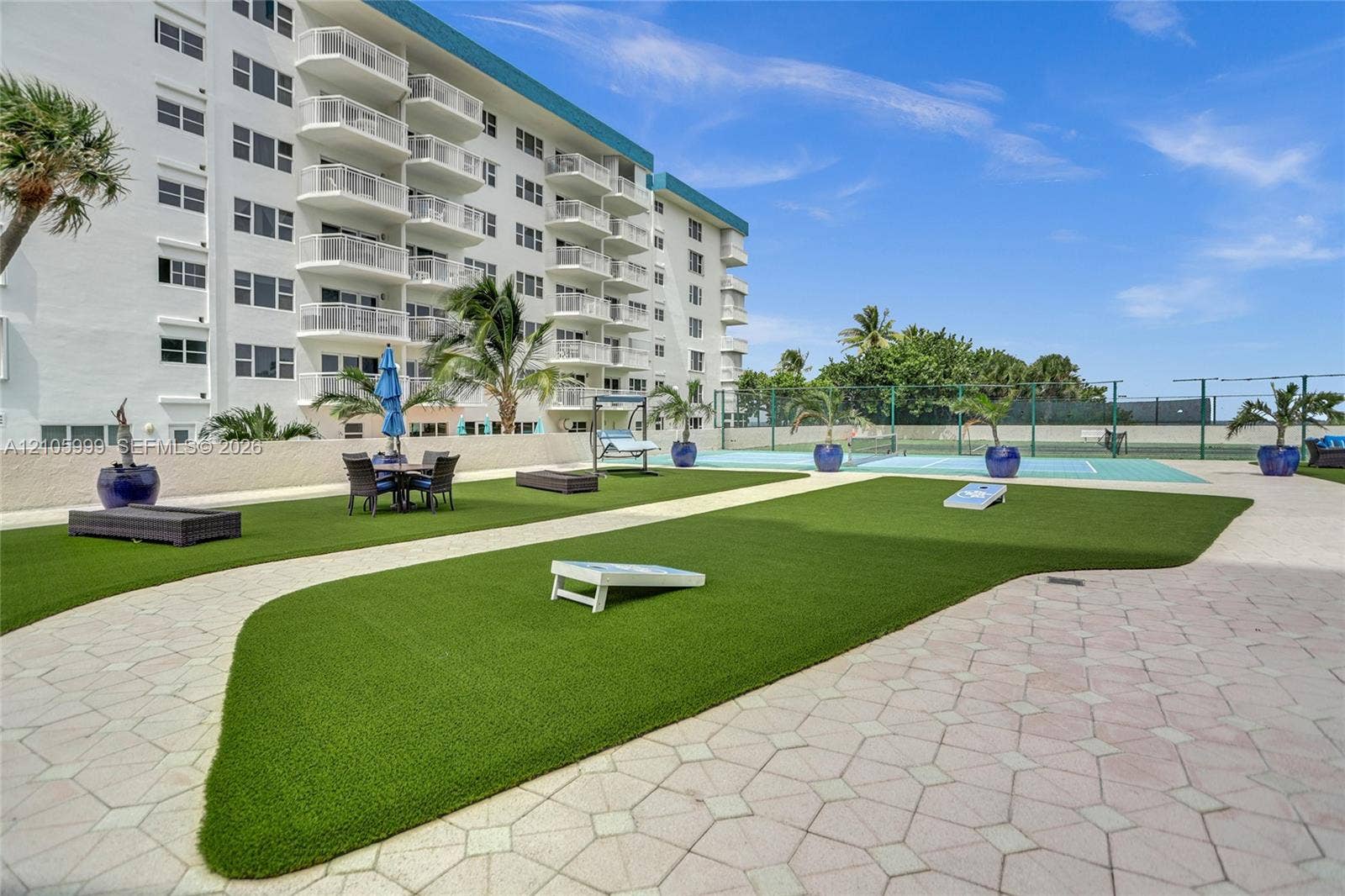 Outdoor recreation area featuring artificial turf, stone-paver hardscaping, a fenced tennis court, and high-rise building with private balconies
