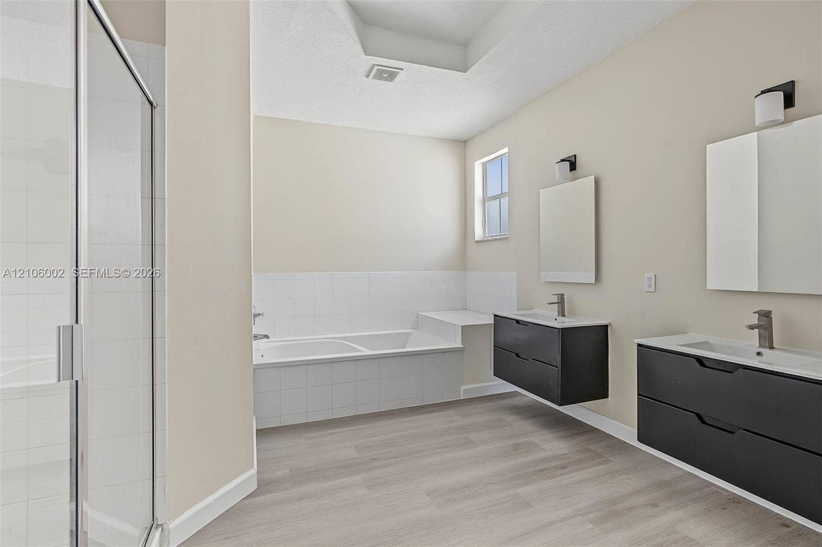 Bathroom featuring dual floating vanities with dark cabinetry, integrated white countertops, rectangular mirrors, and wood-finish flooring, complemented by a tiled soaking tub and a glass-enclosed shower