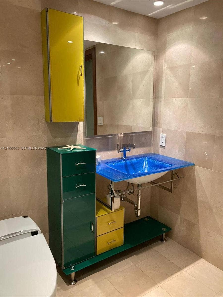 1850 S Ocean Drive, #3309, Hallandale Beach, Hallandale Beach, FL 33009 Bathroom featuring wall-to-wall light-toned stone tiling, a custom vanity with multi-colored high-gloss cabinetry, a blue glass basin sink, and a wall-mounted mirror