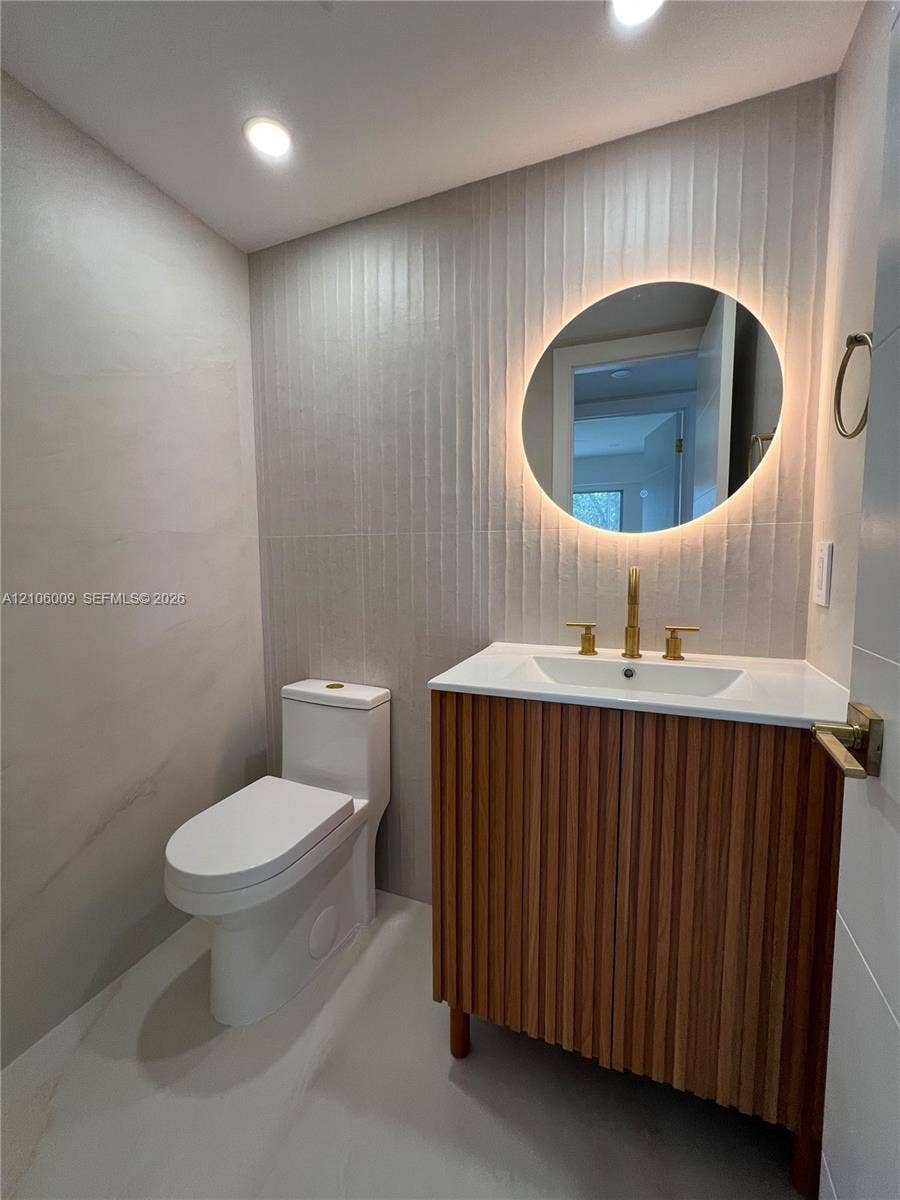 495 Fanshaw L, Boca Raton, FL 33434 Modern vanity with wood-finish vertical slats, white countertop, integrated sink, and brass-finish faucet, complemented by a backlit circular mirror, textured vertical wall tile, and recessed lighting
