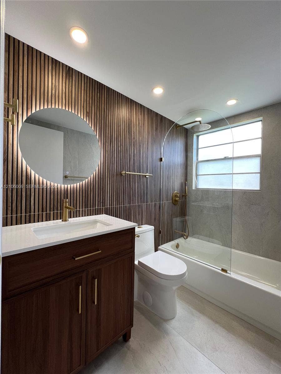 495 Fanshaw L, Boca Raton, FL 33434 Bathroom featuring a wood-slat accent wall, circular LED-backlit mirror, brass-finish fixtures, wooden vanity with a solid-surface countertop, and a glass-enclosed tub with stone-finish tile surround