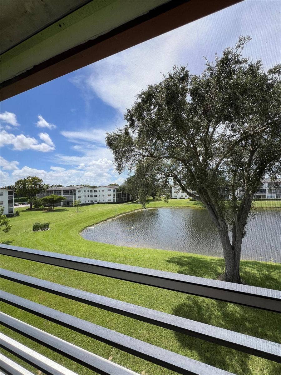 495 Fanshaw L, Boca Raton, FL 33434 Covered balcony with a metal railing overlooking a landscaped green space featuring a pond and mature trees