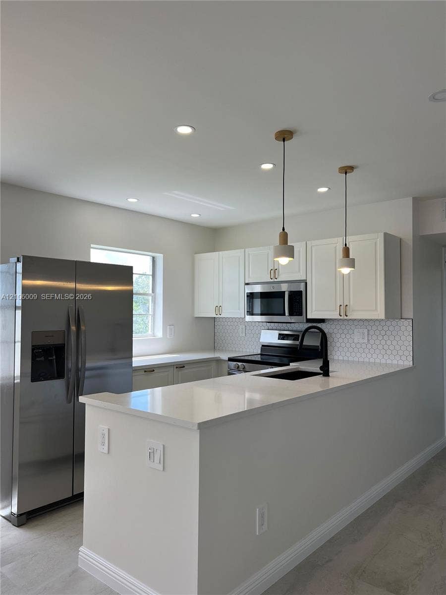 495 Fanshaw L, Boca Raton, FL 33434 Kitchen featuring white shaker-style cabinetry, white solid-surface countertops, stainless steel appliances, a hexagonal tile backsplash, and two minimalist pendant lights