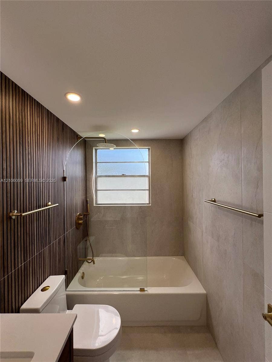 495 Fanshaw L, Boca Raton, FL 33434 Bathtub featuring a frameless glass panel, vertical wood-slat feature wall, large-format tile surround, recessed lighting, and brass-finish plumbing fixtures