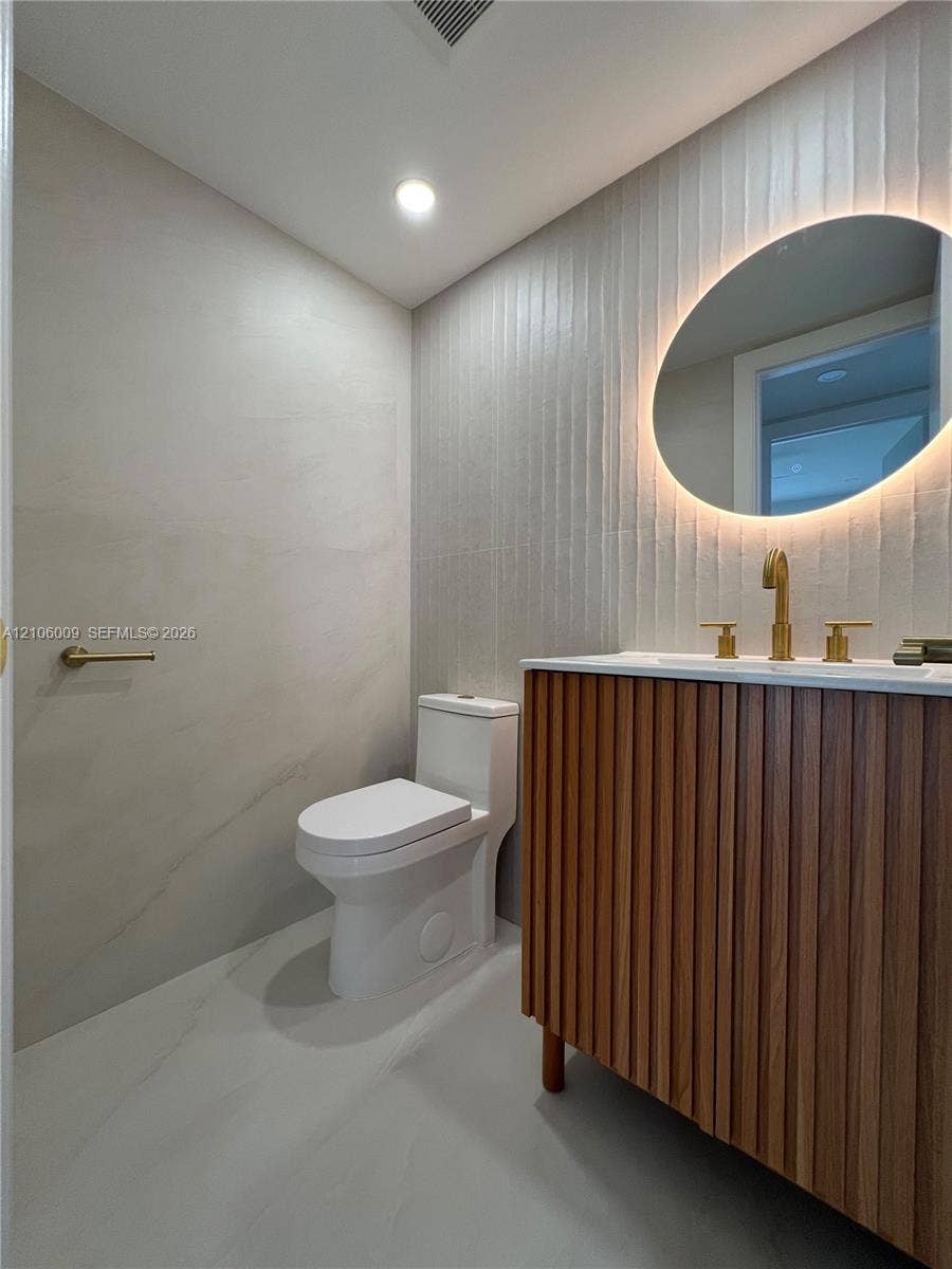 495 Fanshaw L, Boca Raton, FL 33434 Floating vanity with a fluted wood-finish exterior, wall-mounted oval mirror with integrated LED lighting, brushed gold hardware, textured vertical tile accent wall, and large-format neutral floor tiling