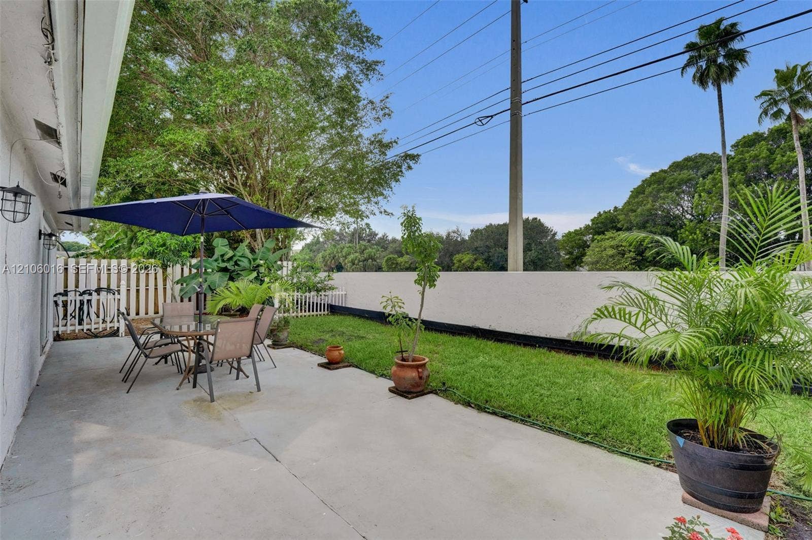 631 NW 135th Terrace, Plantation, FL 33325 Concrete patio featuring a grass lawn, perimeter fencing, wall-mounted exterior lighting, and mature landscaping