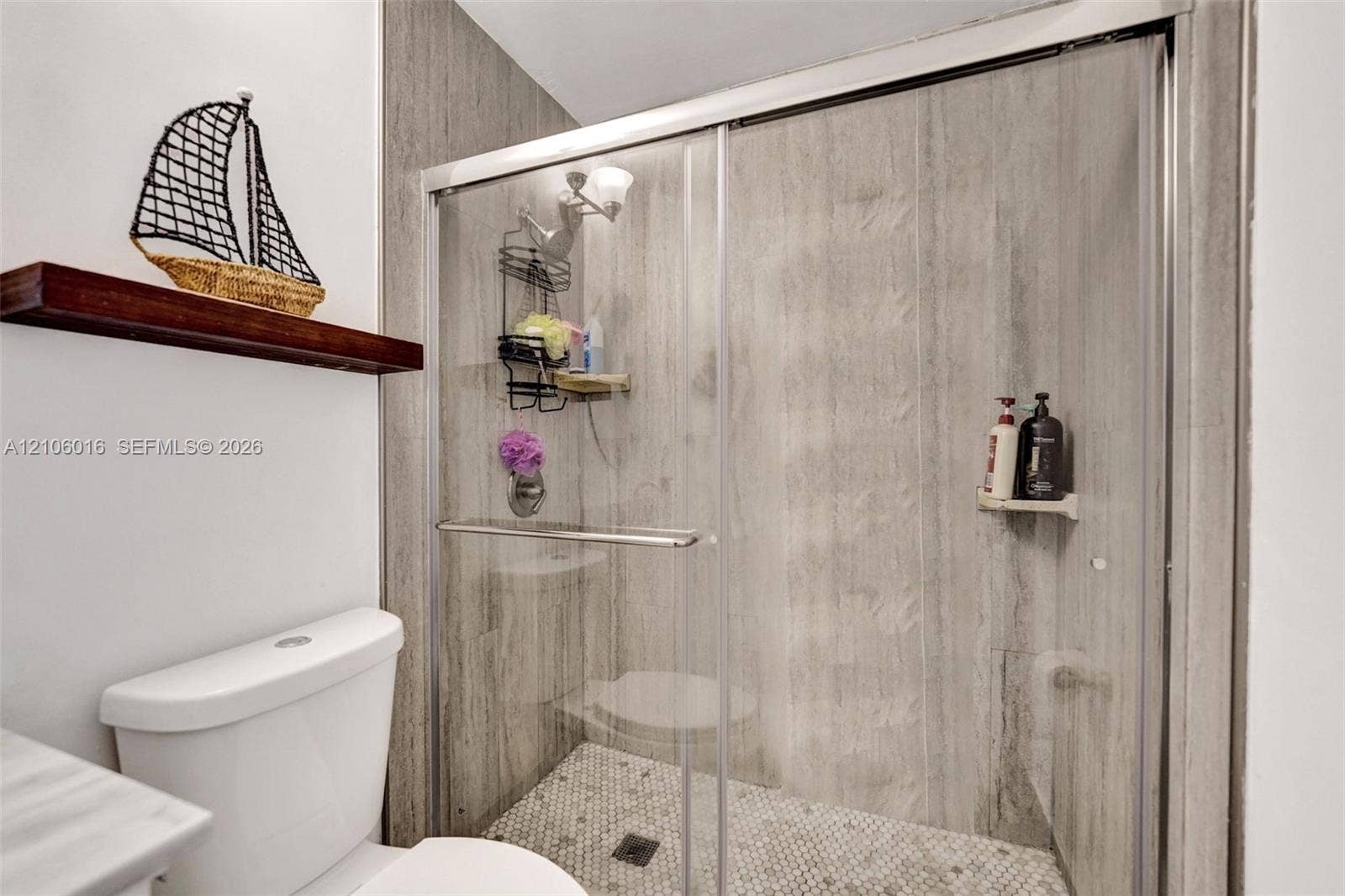 631 NW 135th Terrace, Plantation, FL 33325 Walk-in shower featuring large-format stone-look wall tile, hexagonal mosaic floor tile, and a sliding glass enclosure with a metallic frame, accompanied by a mounted wood-finish display shelf
