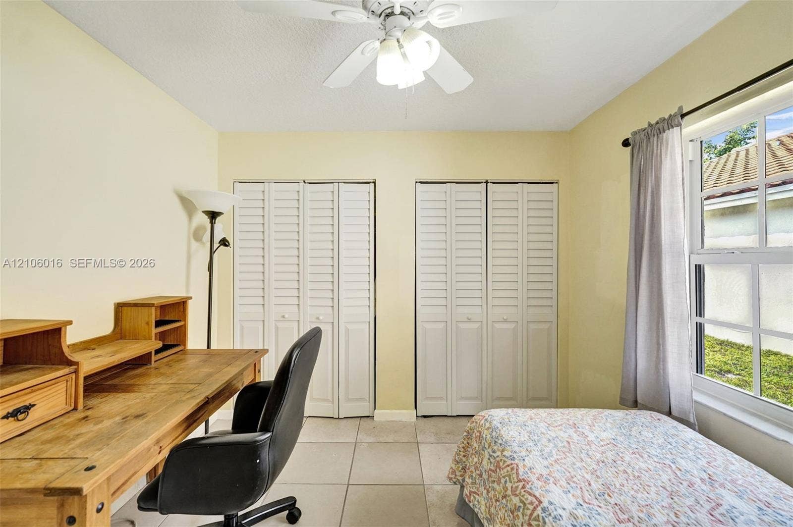 631 NW 135th Terrace, Plantation, FL 33325 Interior room featuring light-toned ceramic tile flooring, two sets of white louvered-style bi-fold closet doors, a multi-blade ceiling fan with integrated lighting, and a single-hung window with a metal rod and drapery hardware