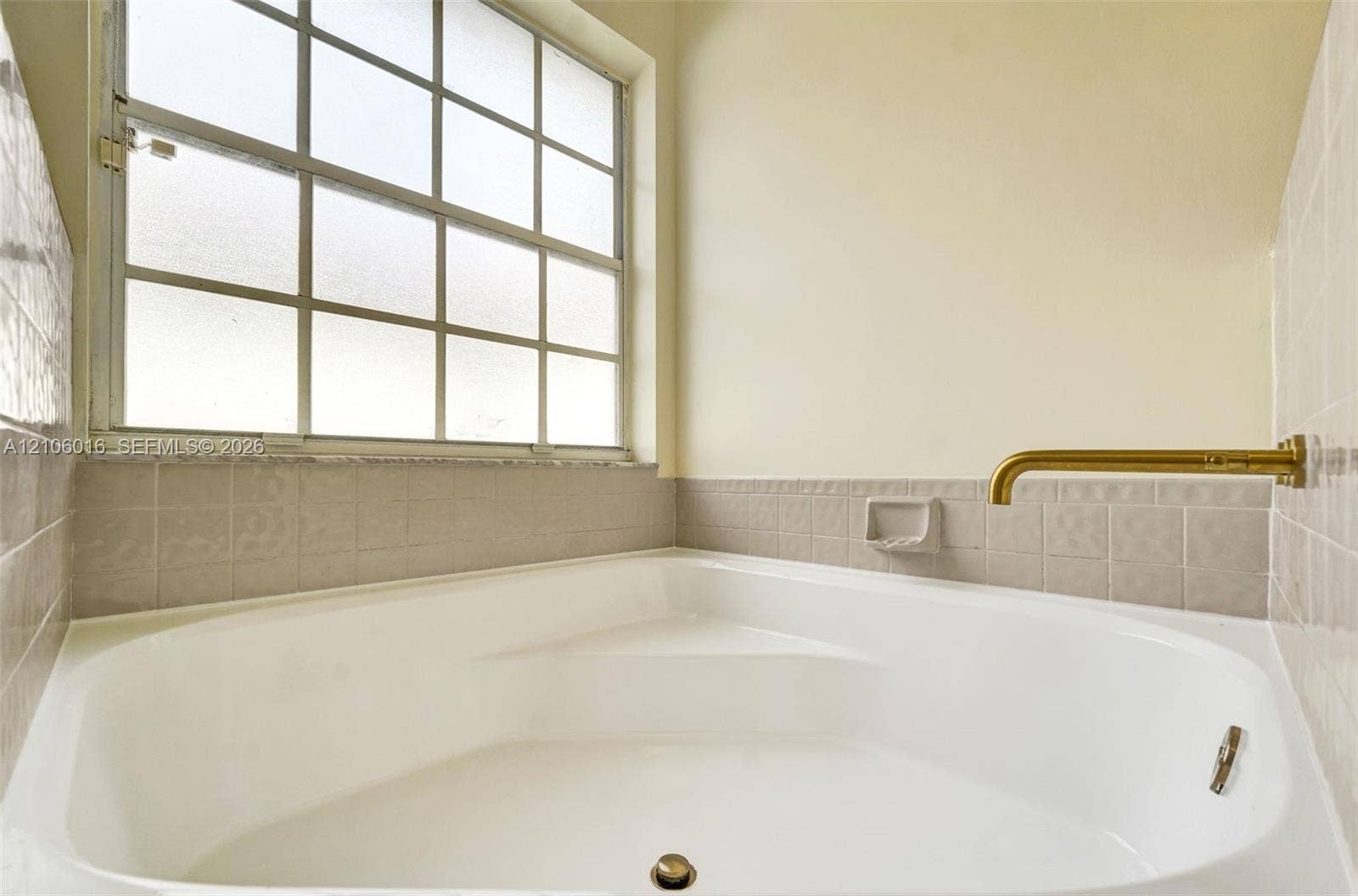 631 NW 135th Terrace, Plantation, FL 33325 Corner soaking tub featuring a metallic wall-mounted faucet, ceramic tile surround, and a multi-pane grid window