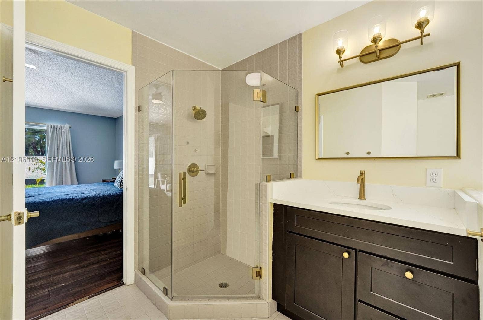 631 NW 135th Terrace, Plantation, FL 33325 Bathroom featuring a corner glass-enclosed shower, dark wood-finish vanity with a light stone-style countertop, rectangular mirror with gold-tone frame, and matching gold-tone plumbing fixtures