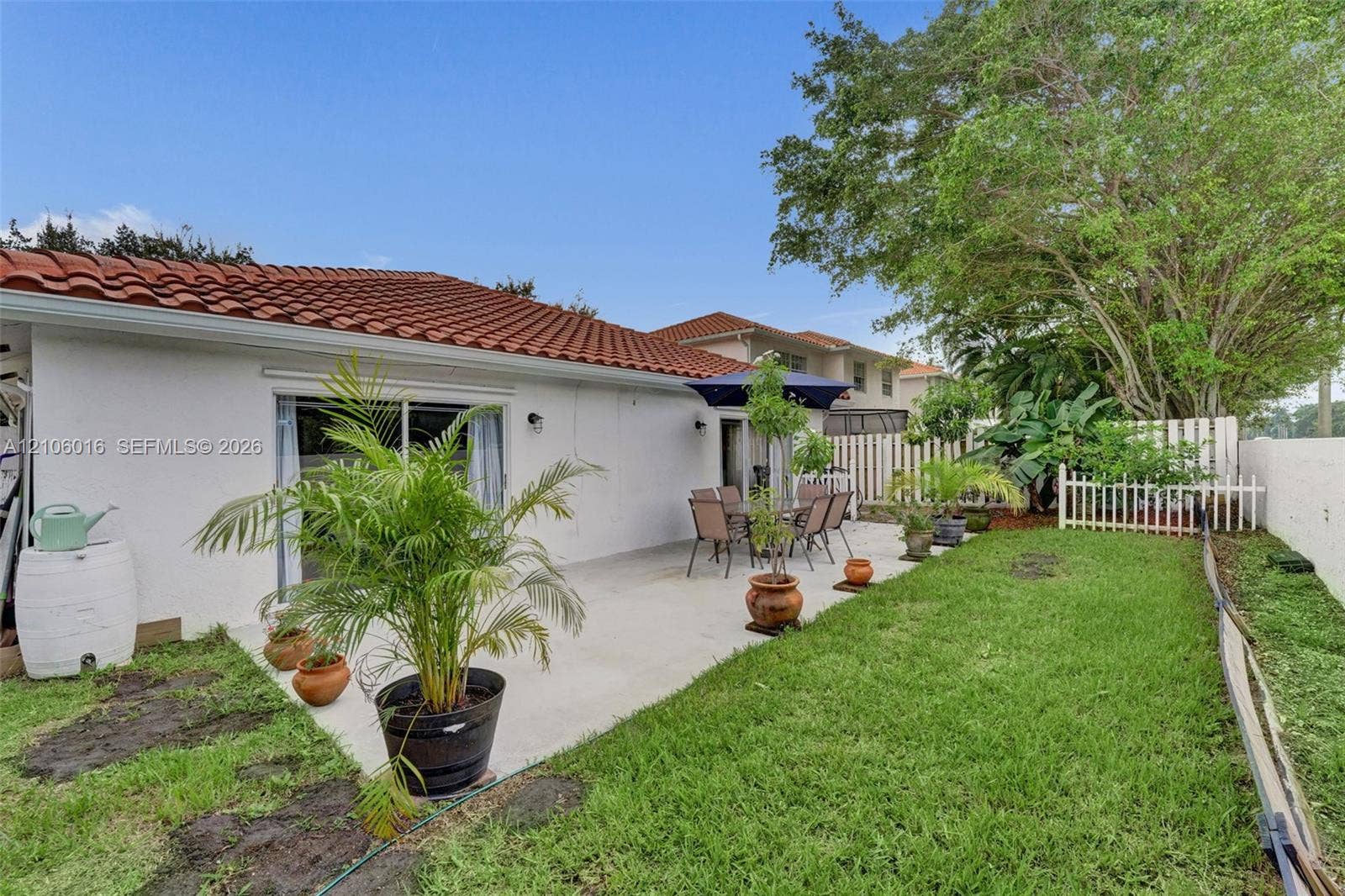 631 NW 135th Terrace, Plantation, FL 33325 Rear exterior featuring a concrete patio, white stucco siding, a terra-cotta tile roof, and a fenced grass lawn with mature trees