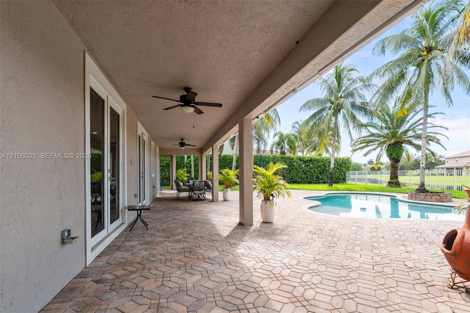 19510 S Coquina Way, Weston, FL 33332 Covered patio featuring paver flooring, recessed ceiling fans, and dual glass-panel doors, leading to a pool area with a fence enclosure and mature palm landscaping