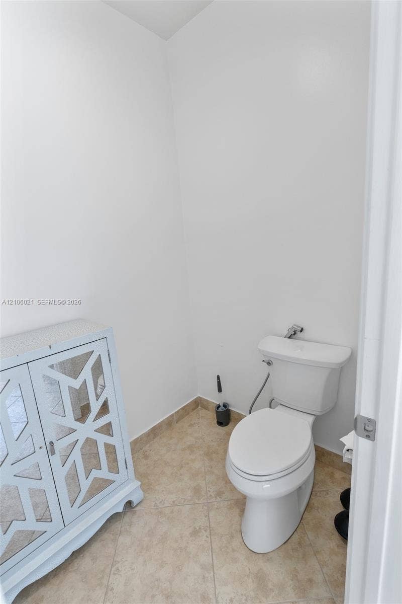 19510 S Coquina Way, Weston, FL 33332 Restroom featuring a white toilet with a wall-mounted bidet sprayer, light-toned ceramic tile flooring, and a freestanding storage cabinet with lattice-paneled doors