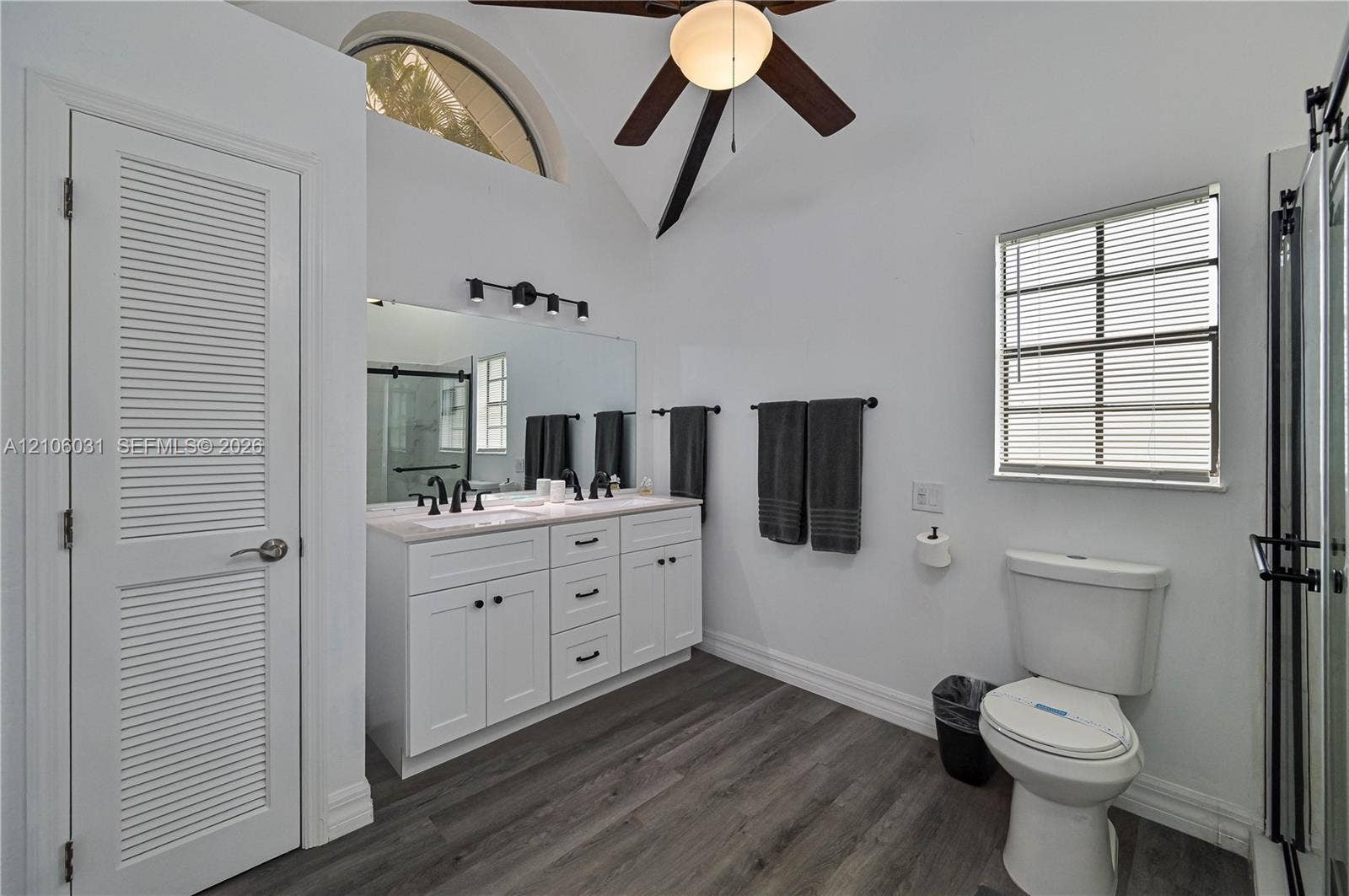 1106 SE 23rd Place, Cape Coral, FL 33990 Double vanity with white cabinetry, neutral stone-finish countertop, dual under-mount sinks, and matte black hardware, featuring wood-finish flooring, vaulted ceilings, and a fixed arched window