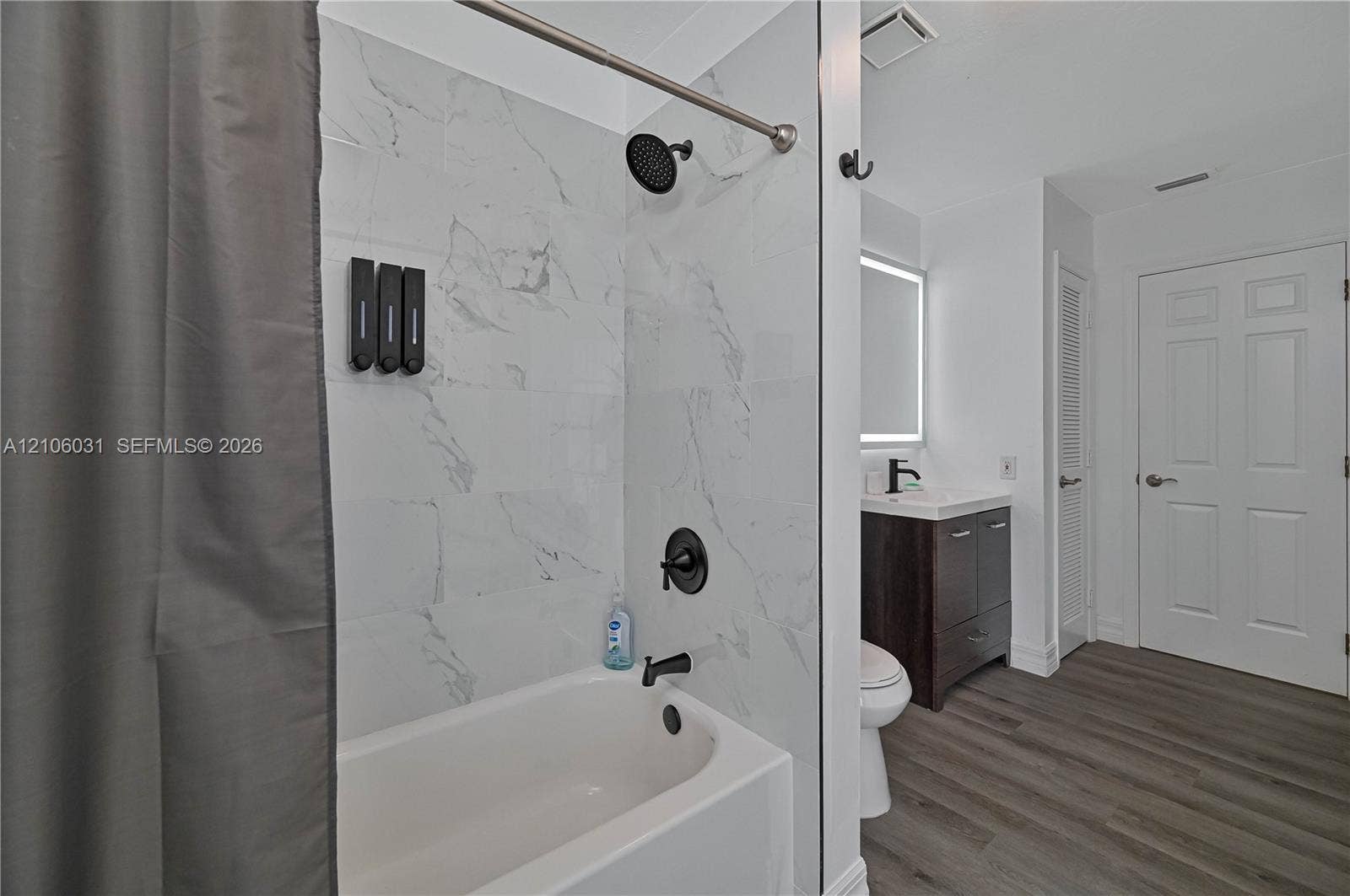 1106 SE 23rd Place, Cape Coral, FL 33990 Bathroom featuring a tub with large-format light-toned tile surround, matte black shower fixtures, wood-finish vanity with a rectangular sink, illuminated mirror, and wood-finish flooring