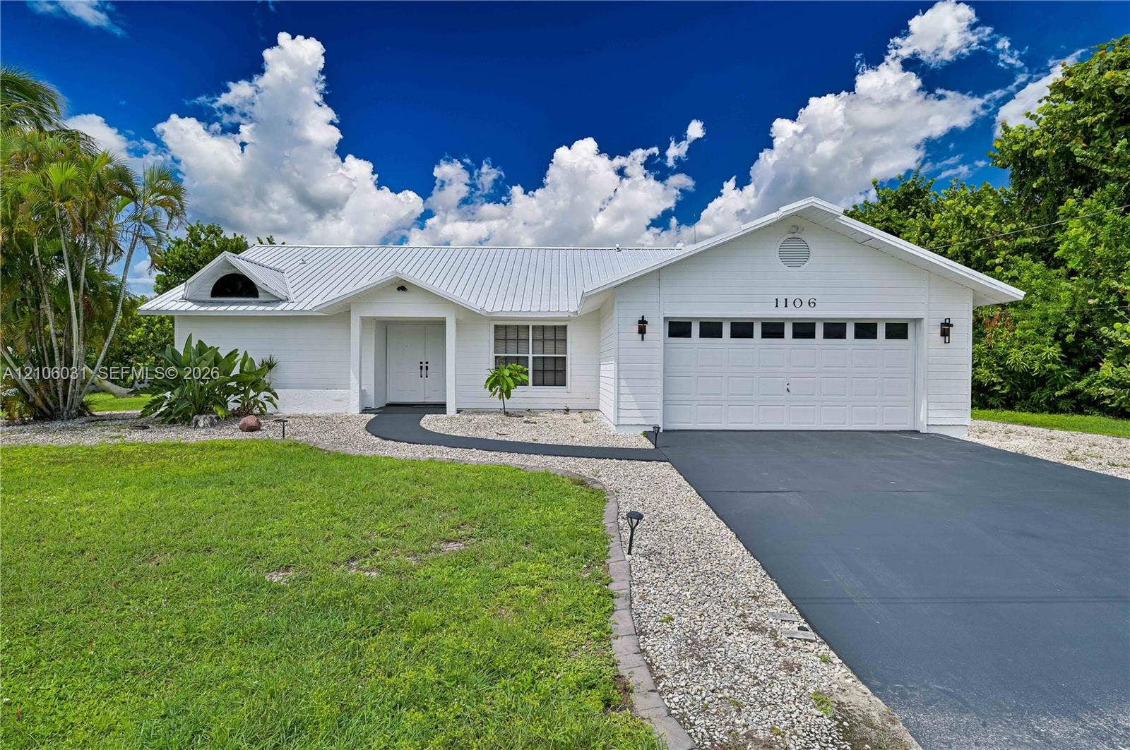 1106 SE 23rd Place, Cape Coral, FL 33990 White exterior siding featuring a metal roof, front-facing two-car garage, and landscaped yard
