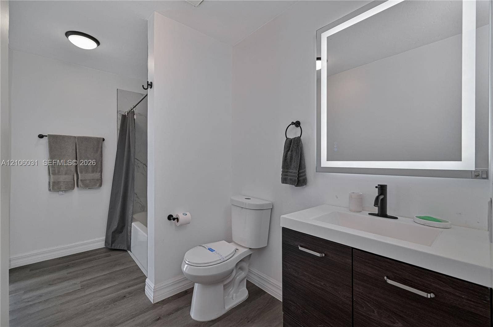 1106 SE 23rd Place, Cape Coral, FL 33990 Bathroom featuring a dark wood-finish vanity with a rectangular integrated sink, matte black faucet, illuminated wall mirror, and grey wood-finish flooring