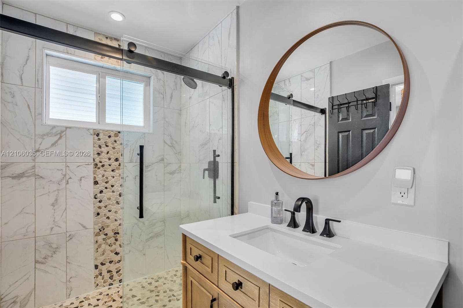 128 Dove Avenue, Tavernier, FL 33070 Bathroom featuring a wooden vanity with a white countertop, circular wood-framed mirror, and a walk-in shower with a sliding glass door, patterned stone accents, and matte black hardware
