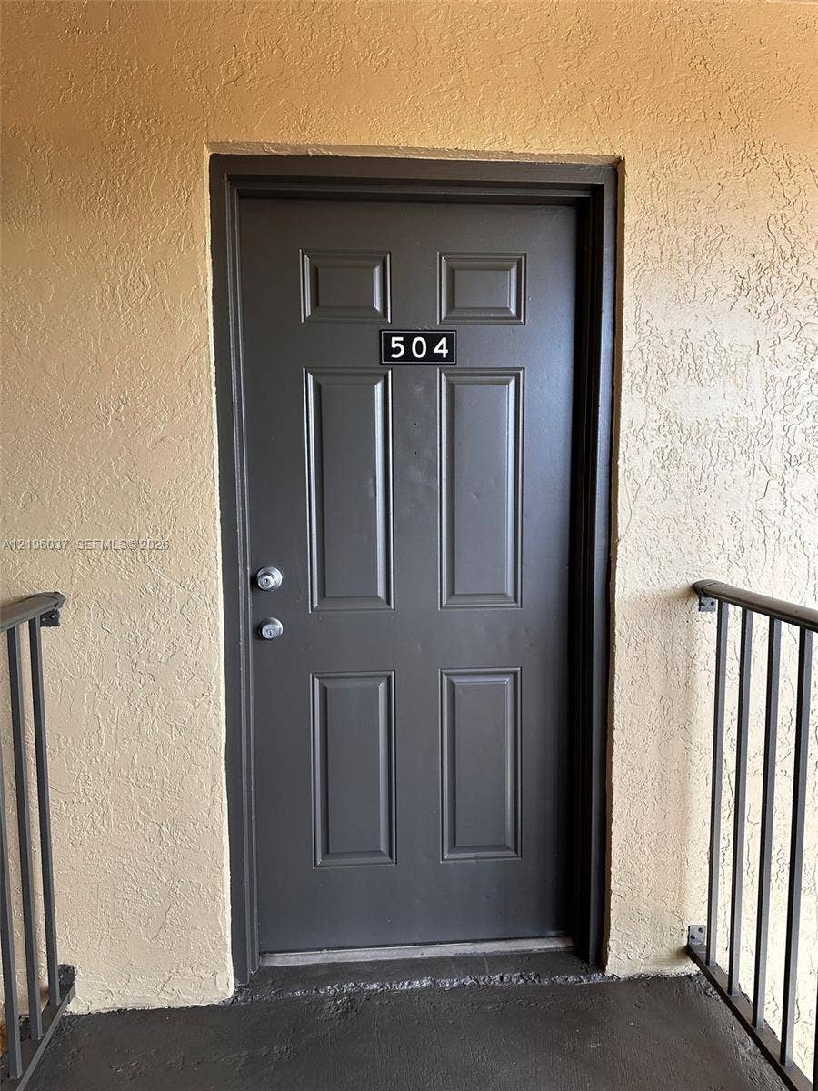 14951 SW 82nd Lane, #17-504, Miami, FL 33193 Six-panel entry door finished in a dark neutral tone, featuring a dual-lock handle set and numerical address plaque, framed by textured exterior stucco walls