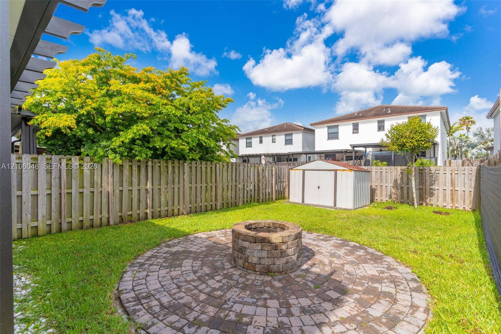 25134 SW 108th Court, Homestead, FL 33032 Backyard featuring a circular paver patio, stone-finish fire pit, wood privacy fencing, and metal storage shed