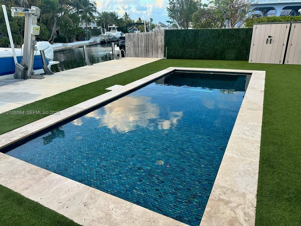 179 Azalea Street, Islamorada, FL 33070 Rectangular pool with tile interior and natural stone coping, synthetic lawn surface, boat lift, concrete bulkhead, and wood-panel privacy fencing