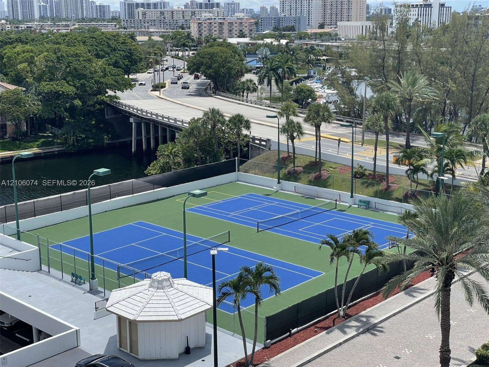 300 Three Islands Boulevard, #308, Hallandale Beach, Hallandale Beach, FL 33009 Dual tennis courts featuring hard-surface playing areas, perimeter fencing, overhead lighting, and a central gazebo