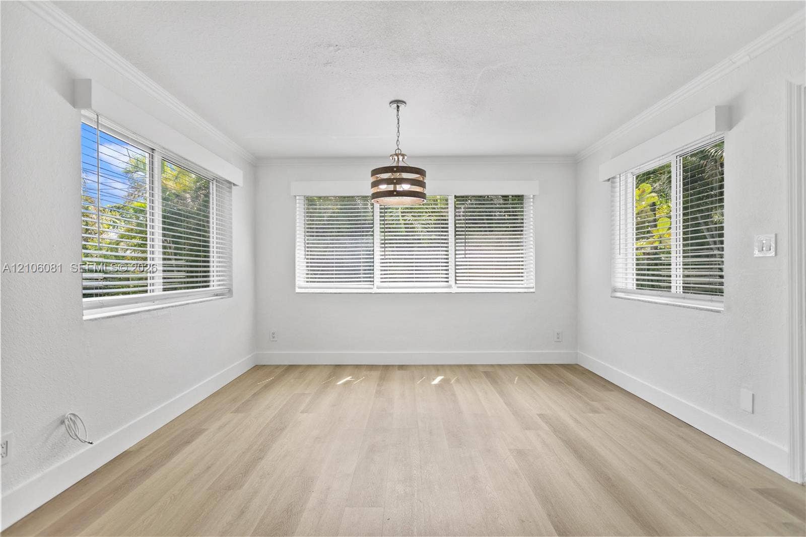 Light-filled room featuring wood-finish flooring, crown molding, a central drum pendant light fixture, and windows equipped with horizontal blinds