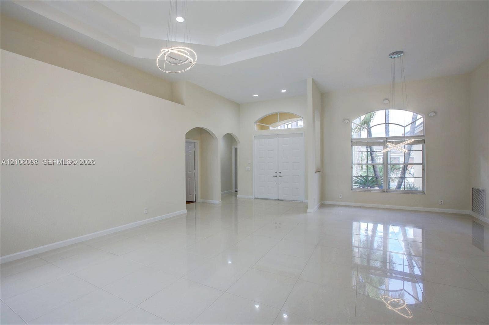 Open-concept interior featuring high tray ceilings, polished tile flooring, arched doorways, a double-entry door with transom window, and an expansive multi-pane arched window