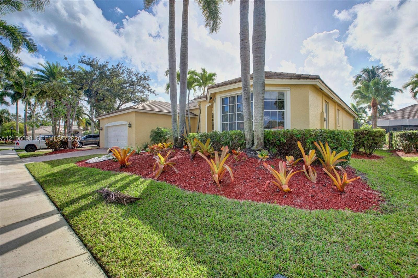 Single-story exterior featuring a stucco facade, multi-pane bay window, attached garage, and landscaped garden beds with palm trees