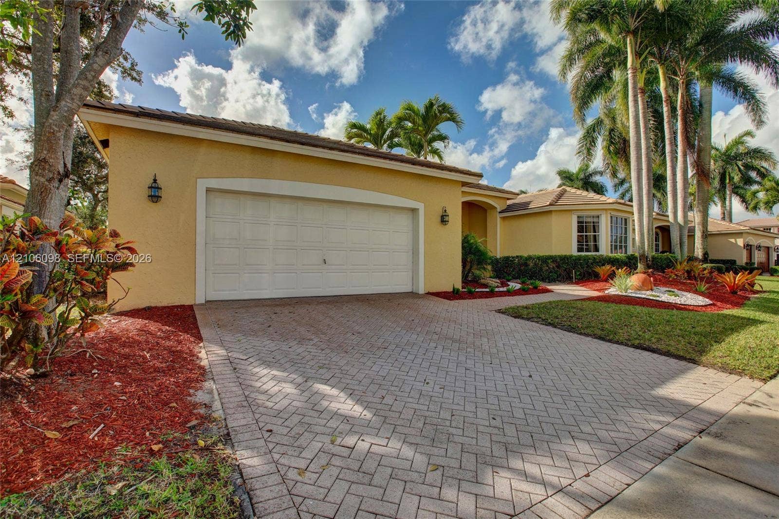 Stucco exterior residence featuring an arched single-bay garage door, herringbone-patterned paver driveway, and mature palm tree landscaping