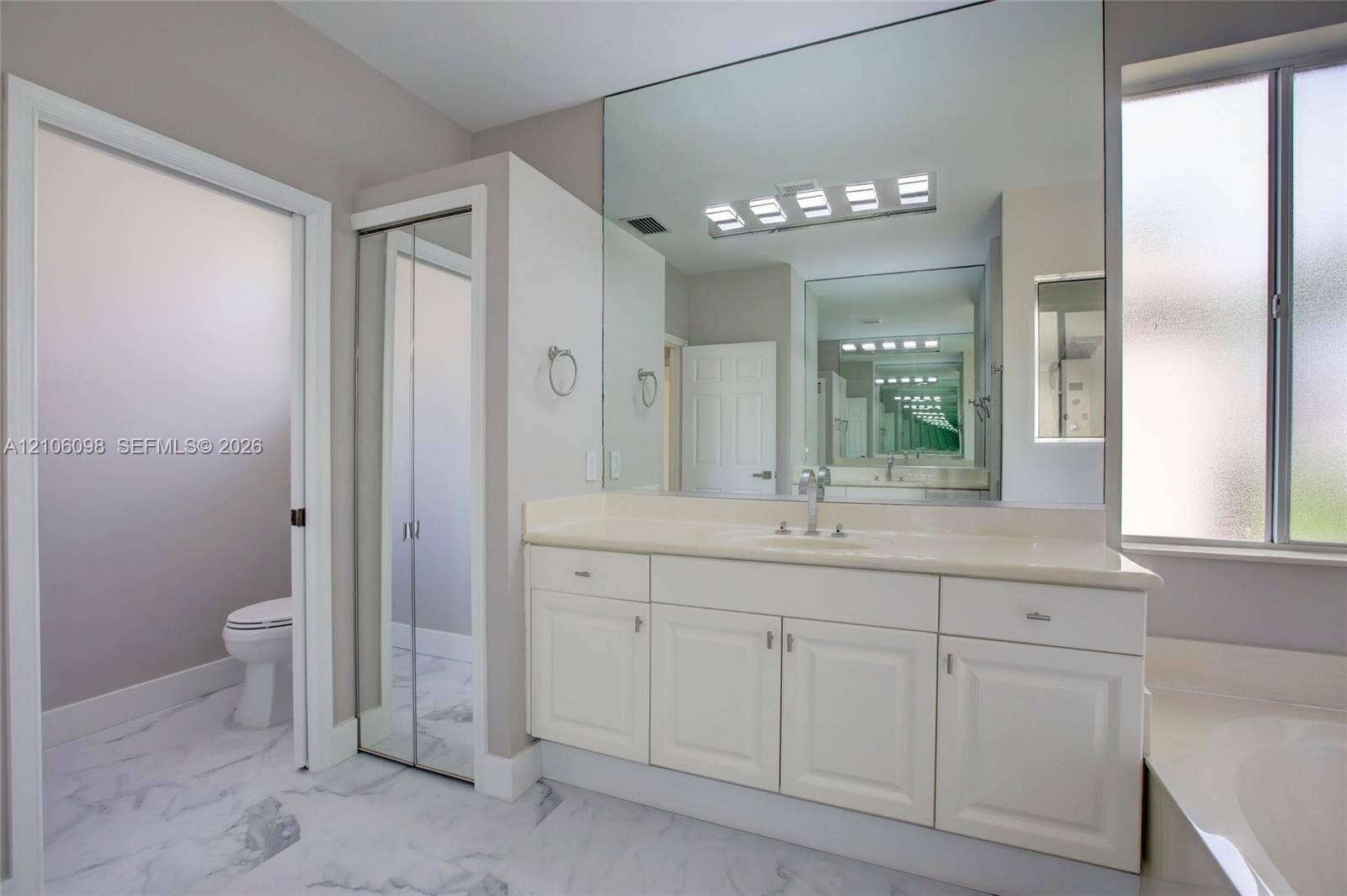 Bathroom featuring white cabinetry, solid-surface vanity countertop, full-width wall mirror, chrome-finish plumbing fixtures, and marble-patterned tile flooring