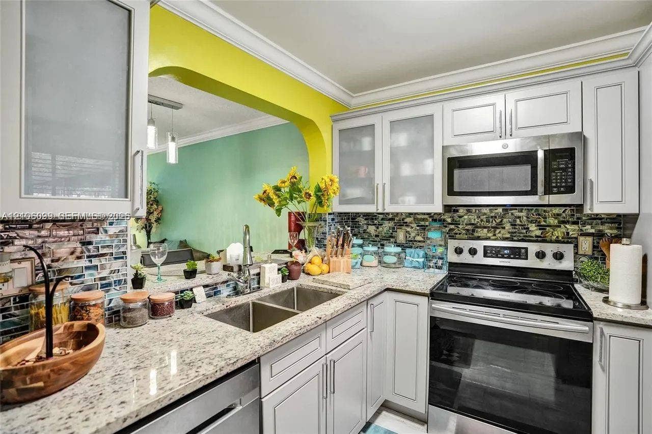 7840 NW 50th Street, #105, Lauderhill, FL 33351 Kitchen featuring shaker-style cabinetry with frosted glass inserts, speckled stone countertops, mosaic tile backsplash, and stainless steel appliances including a built-in microwave