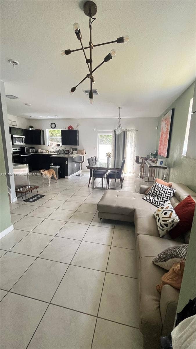 Open-concept interior featuring large-format tile flooring, modern multi-arm chandelier lighting, and a kitchen equipped with dark cabinetry