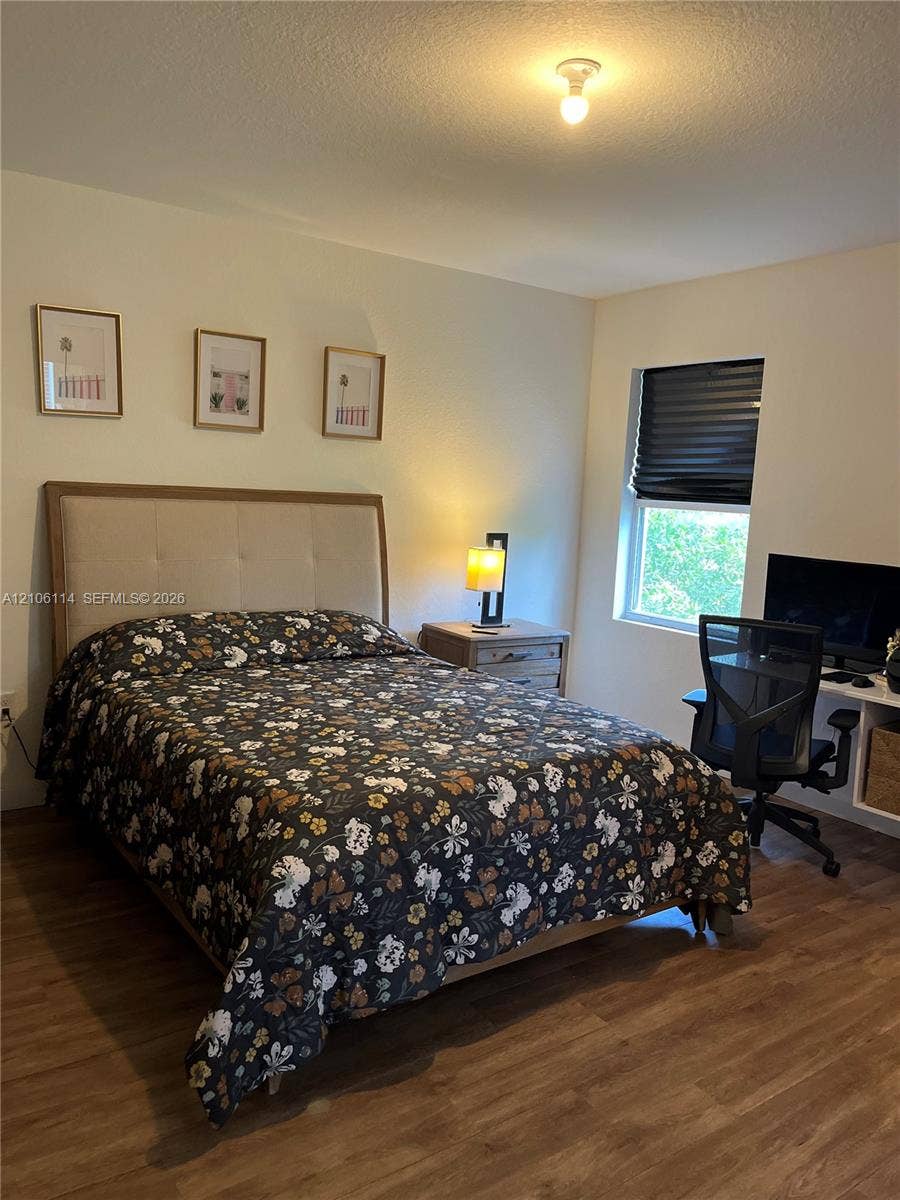 Wood-finish flooring, neutral wall tones, single-light ceiling fixture, window with fitted shade, and built-in desk surface