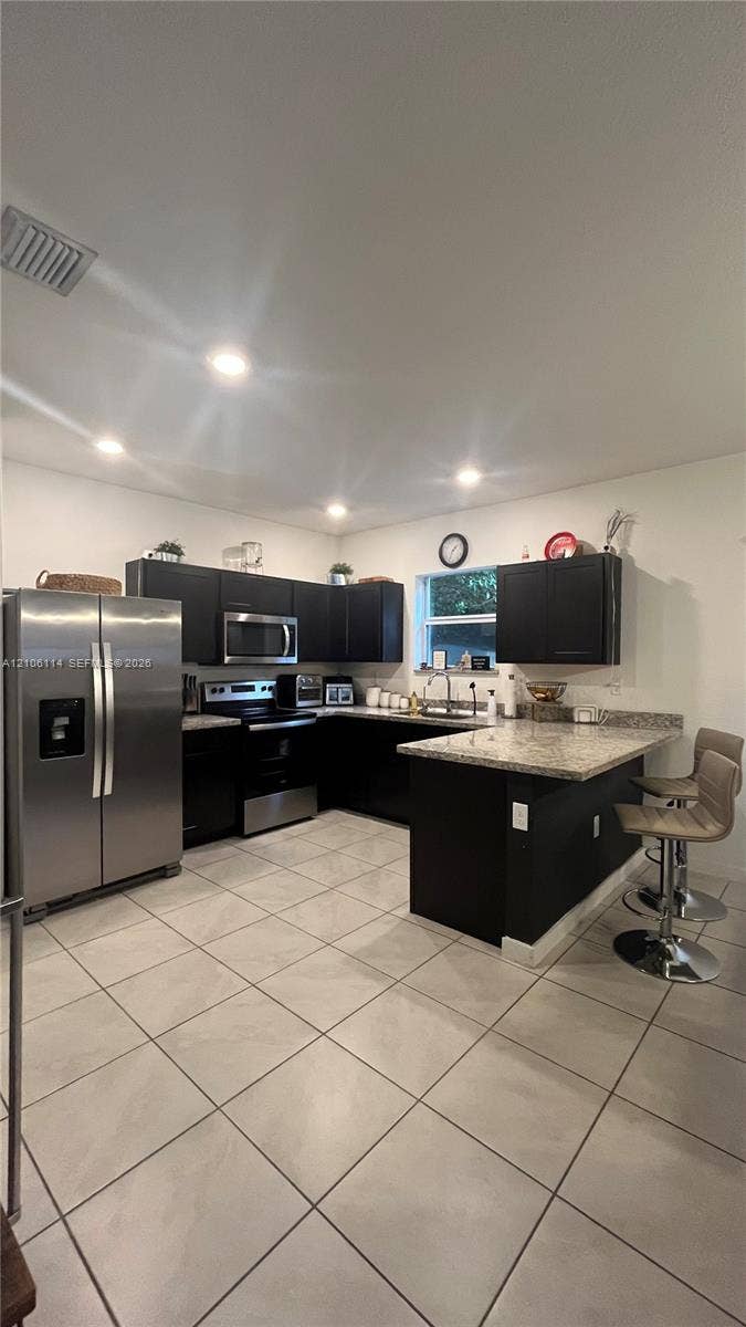 Kitchen featuring dark cabinetry, stone-finish countertops, stainless steel appliances, a breakfast bar, and tile flooring