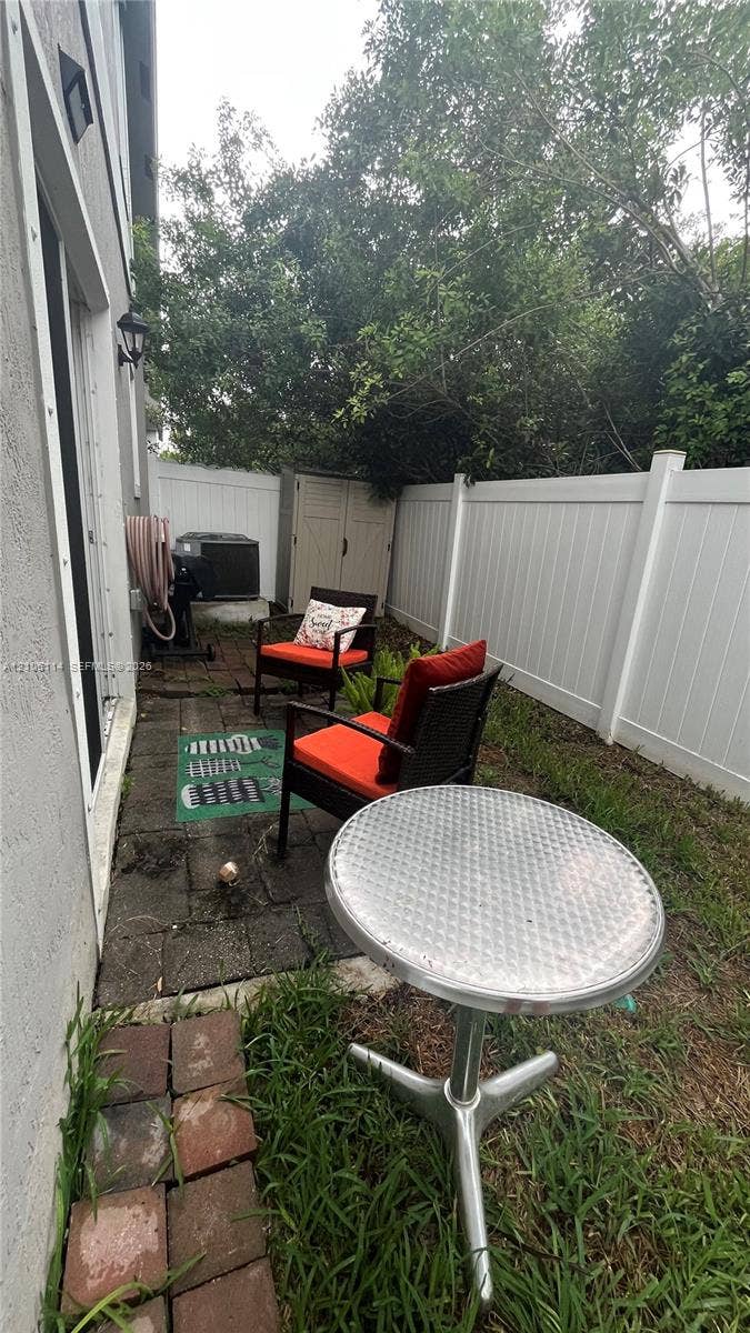 Outdoor area featuring a paved stone patio, white vinyl privacy fencing, and exterior wall-mounted lighting