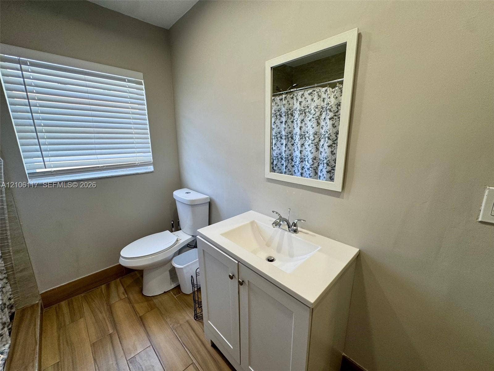 Bathroom featuring a white vanity cabinet with a stone-top basin, wood-finish flooring, a wall-mounted framed mirror, and a window with horizontal blinds