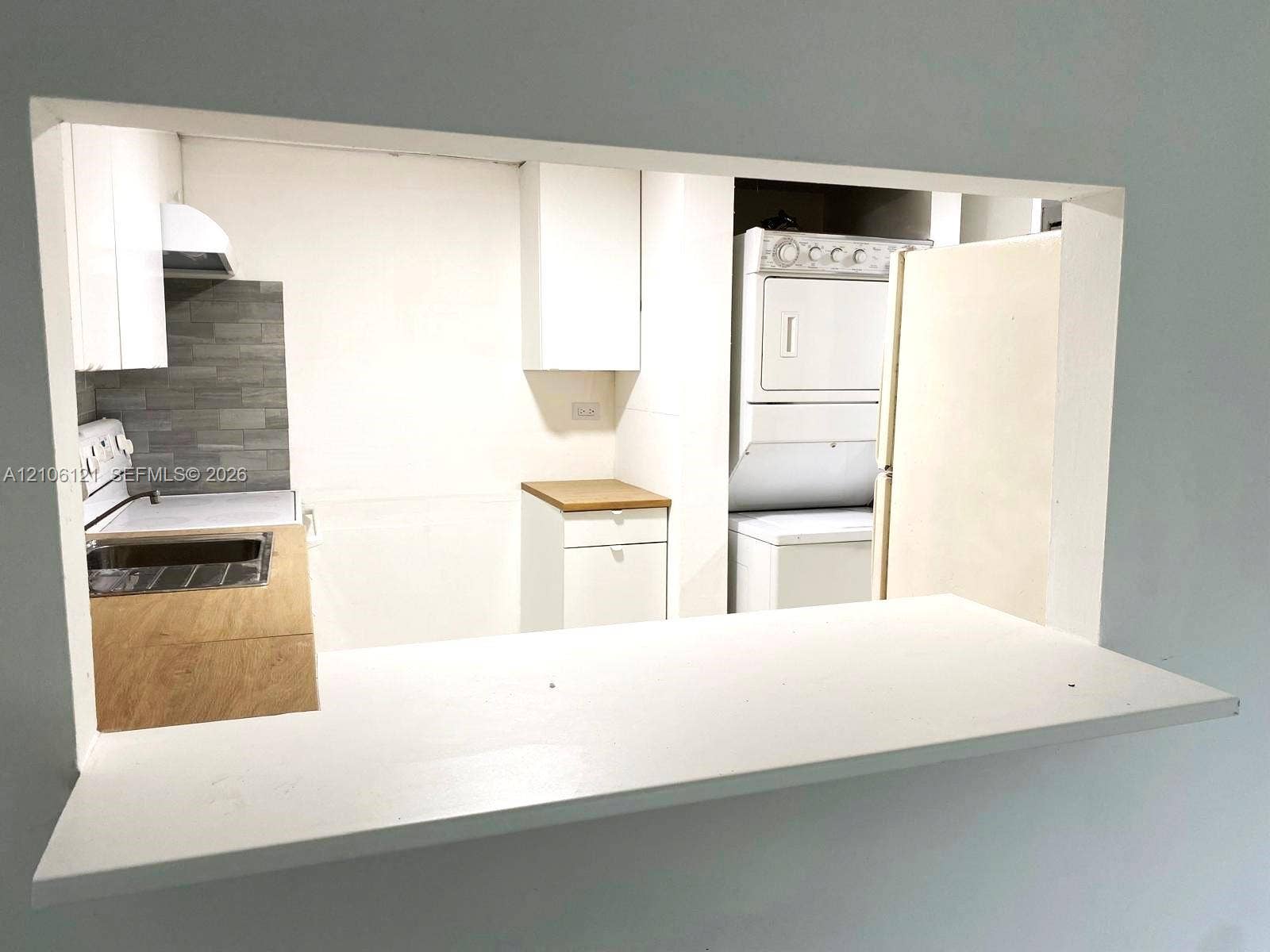 Kitchen pass-through window featuring a white ledge, light wood-finish cabinetry, gray tile backsplash, and a stacked laundry appliance configuration