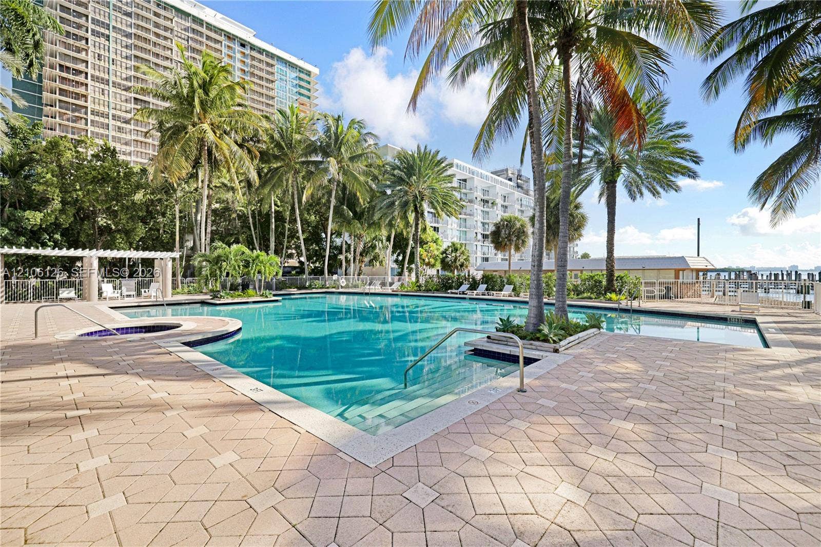 Swimming pool with integrated spa and decorative stone paver decking, surrounded by mature palm trees and a shaded pergola structure