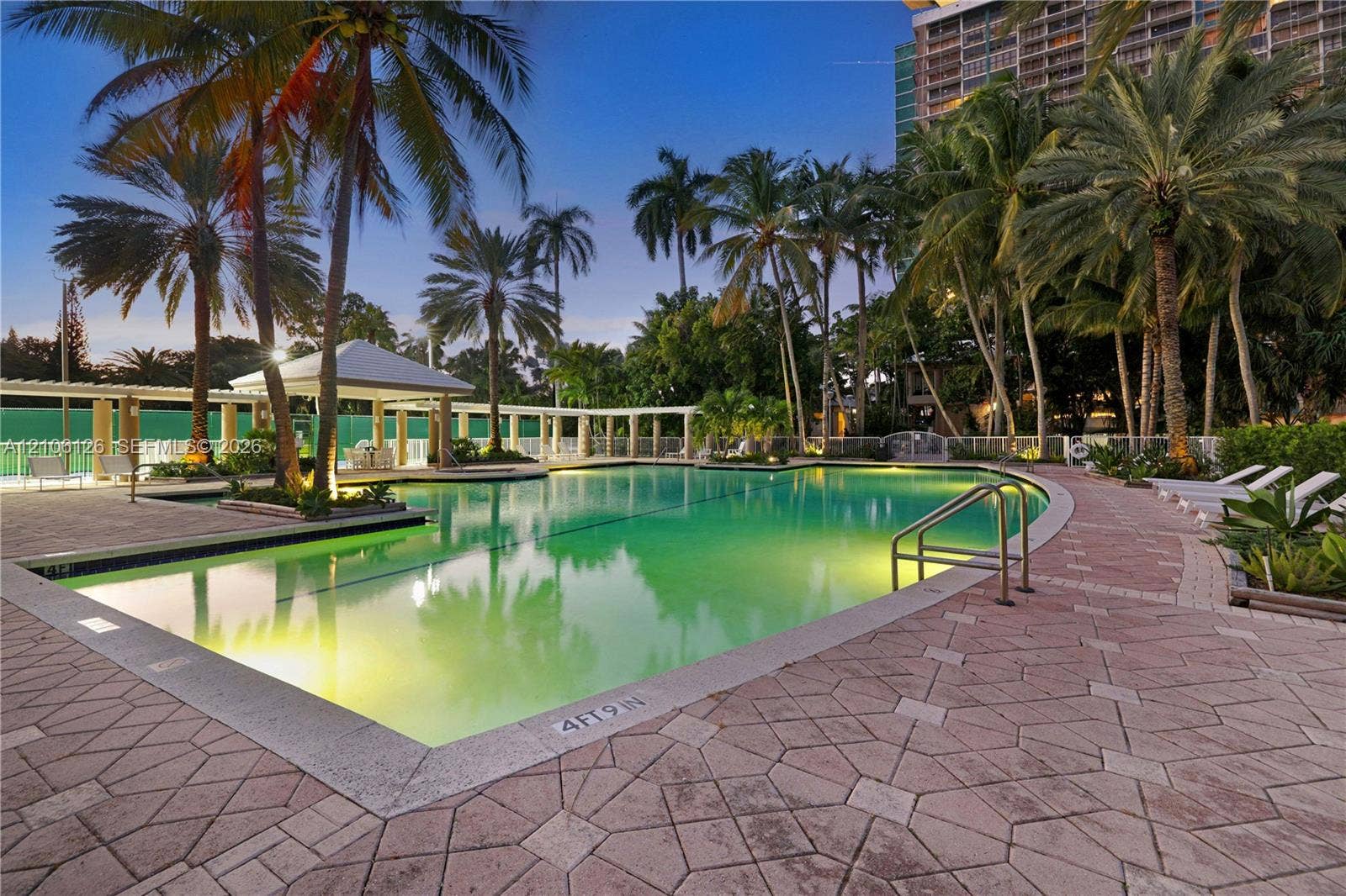 In-ground swimming pool featuring a patterned paver deck, covered gazebo, stainless steel ladder, and mature palm trees