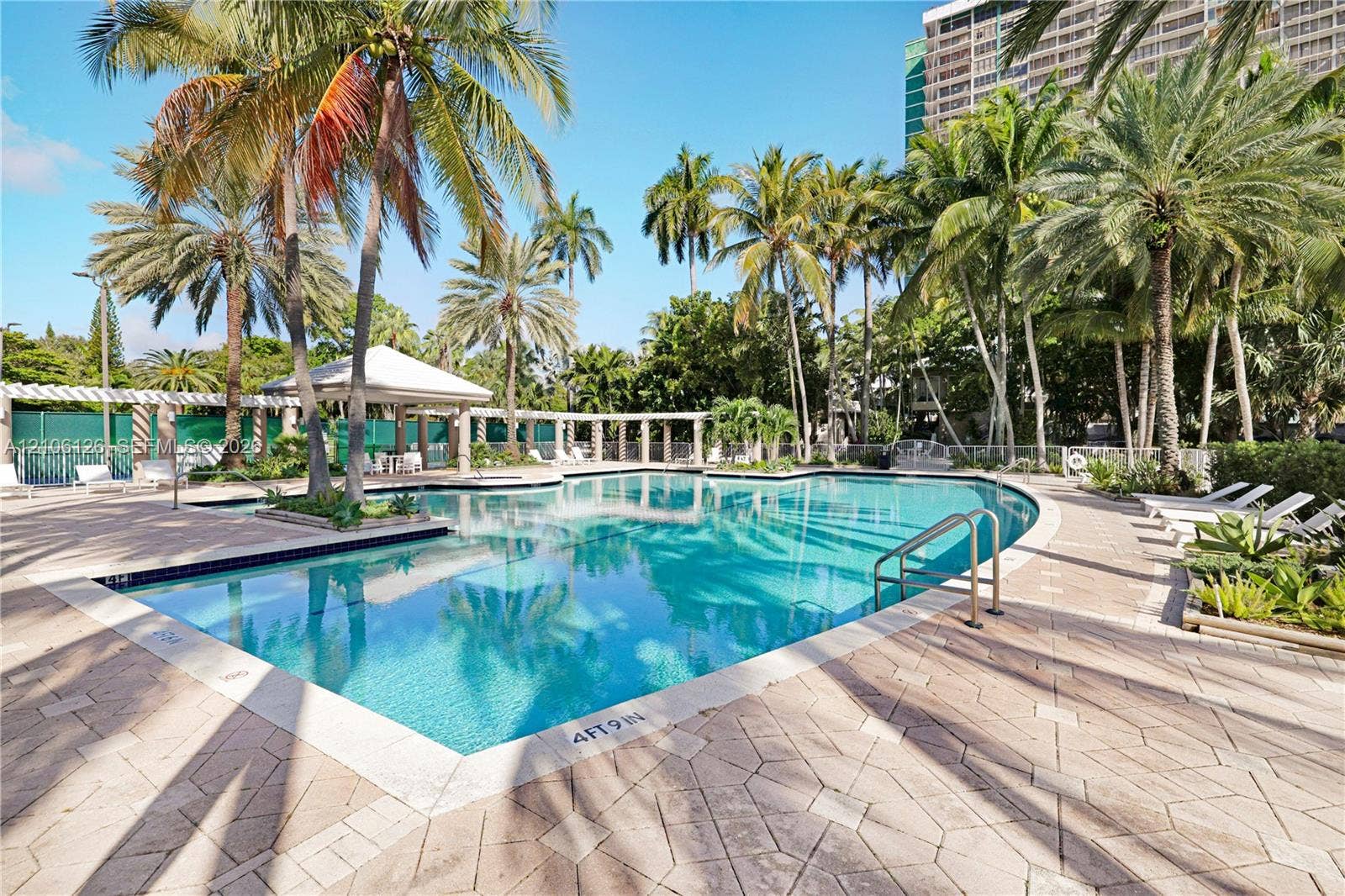 In-ground swimming pool featuring a tiled perimeter, integrated metal handrails, a covered pavilion, and surrounding paver decking lined with mature palm trees
