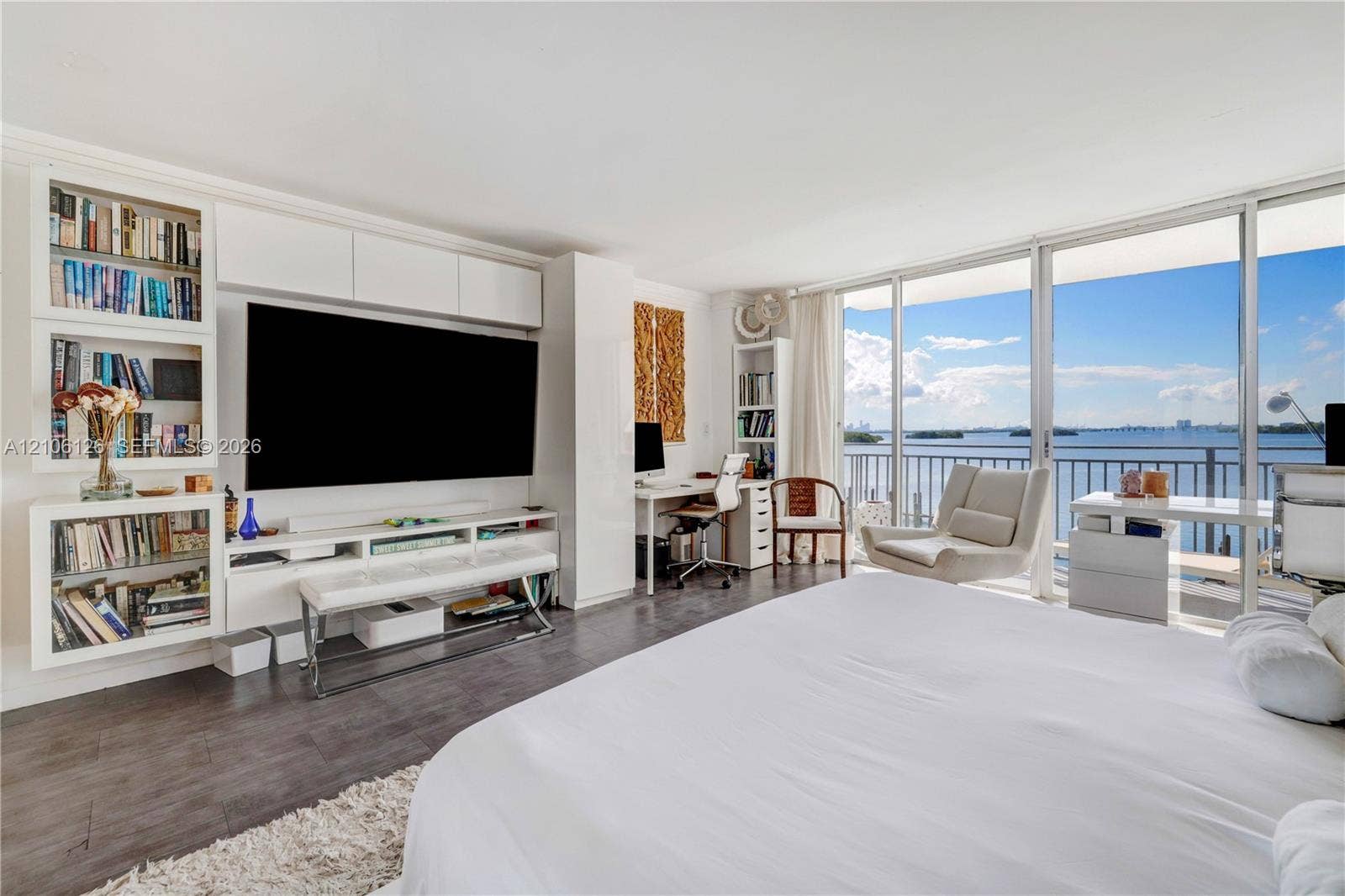 Floor-to-ceiling glass windows, wood-finish flooring, built-in cabinetry with integrated shelving, and a balcony featuring a water backdrop