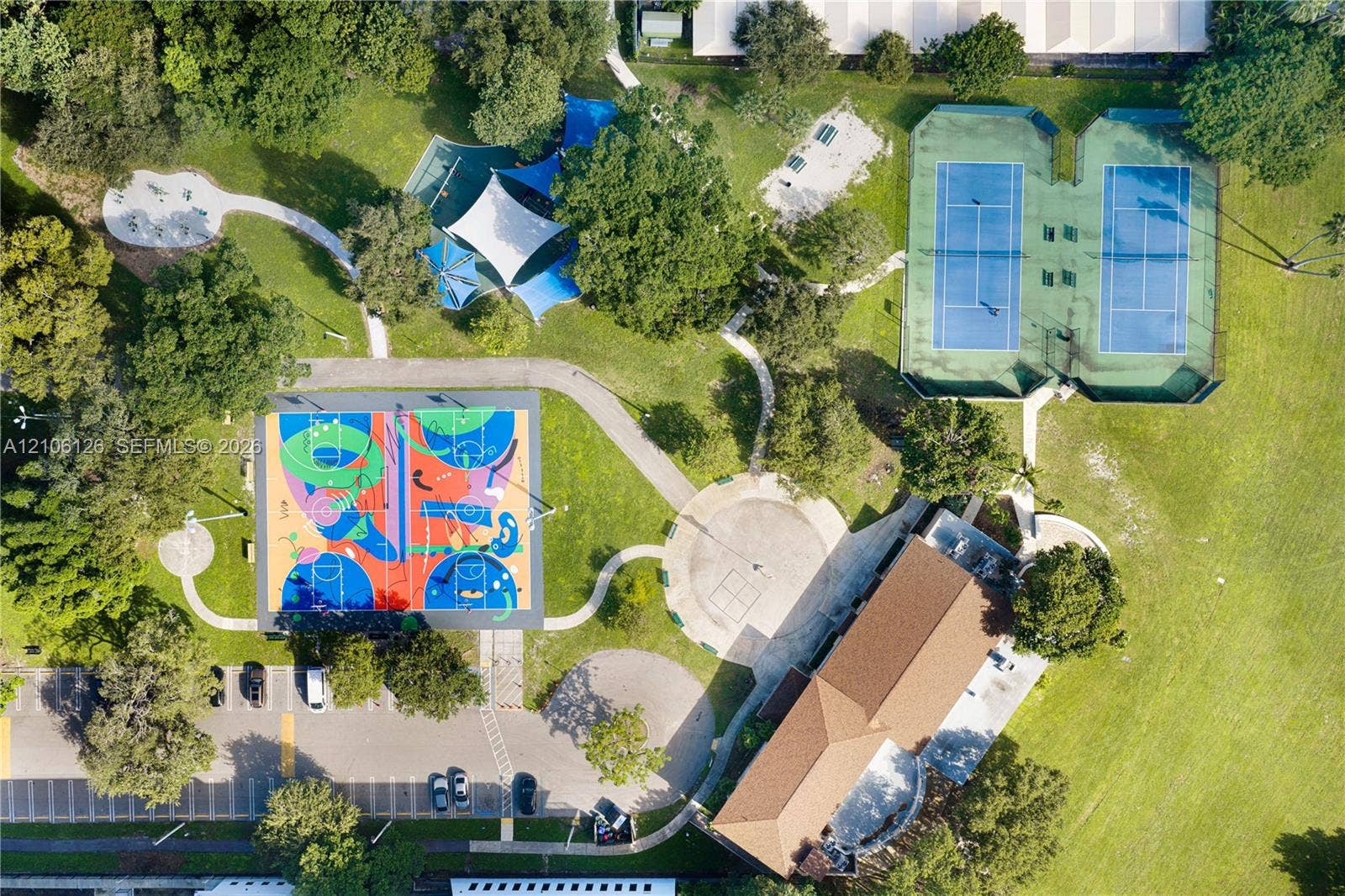 Aerial perspective of community grounds featuring two tennis courts, a multi-use court with sport surfacing, shaded structures, and paved walkways