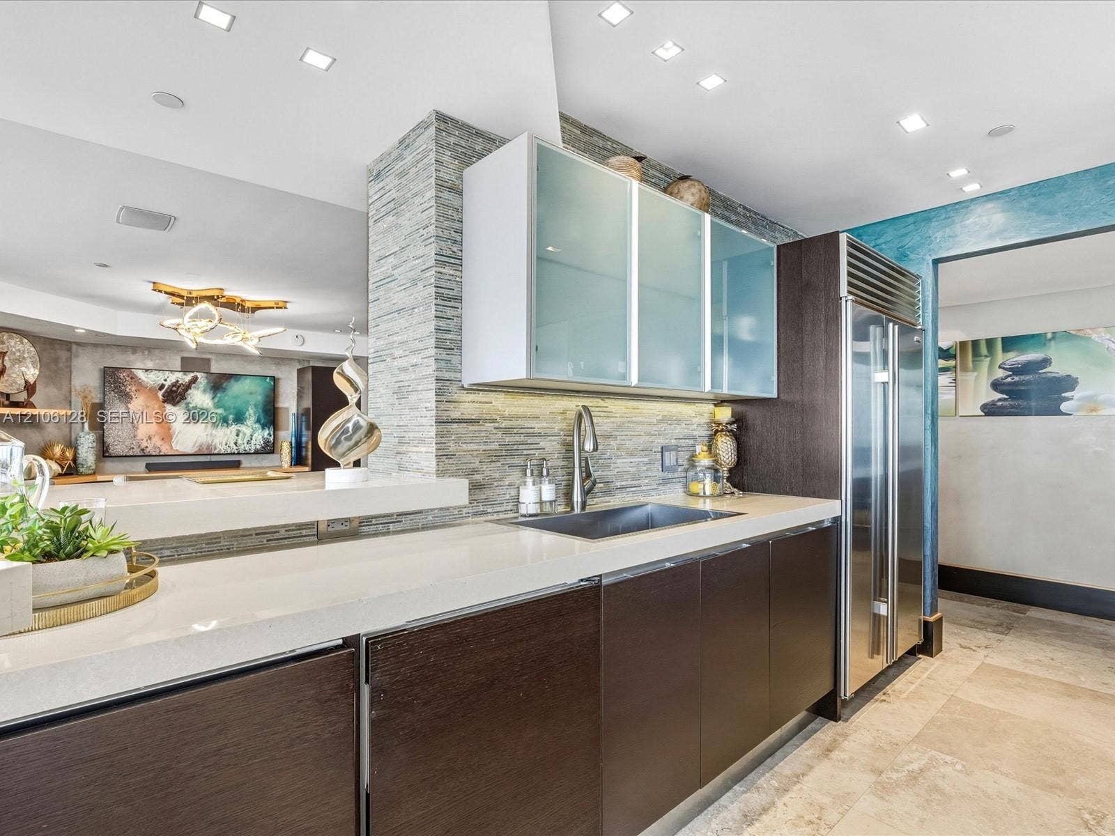 Modern kitchen featuring dark wood-finish cabinetry, frosted glass upper cabinets, a light-toned countertop, a built-in stainless steel refrigerator, and a textured stacked-stone backsplash