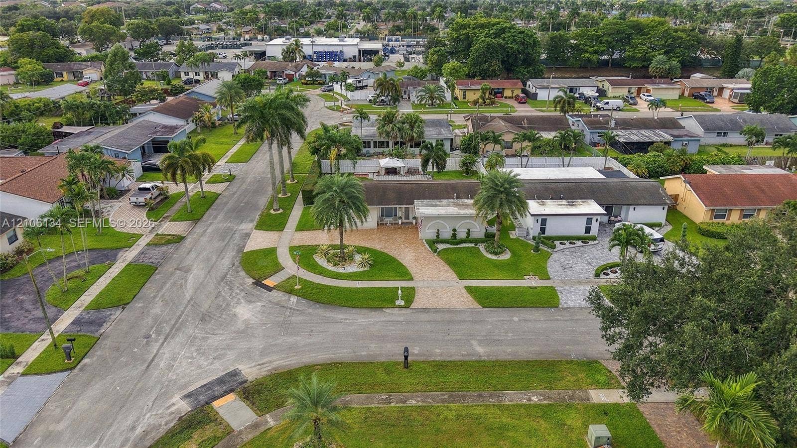 Aerial perspective of a single-story residence featuring a sprawling corner lot, multi-car paver driveway, manicured landscaping, and mature palm trees