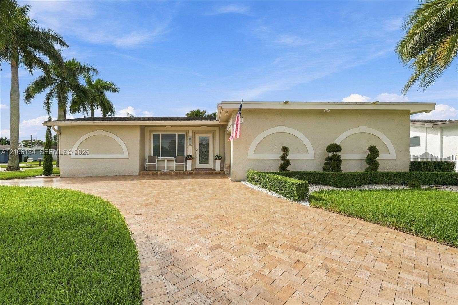 Single-story exterior featuring a stone-paver driveway, decorative arched stucco detailing, and mature palm tree landscaping