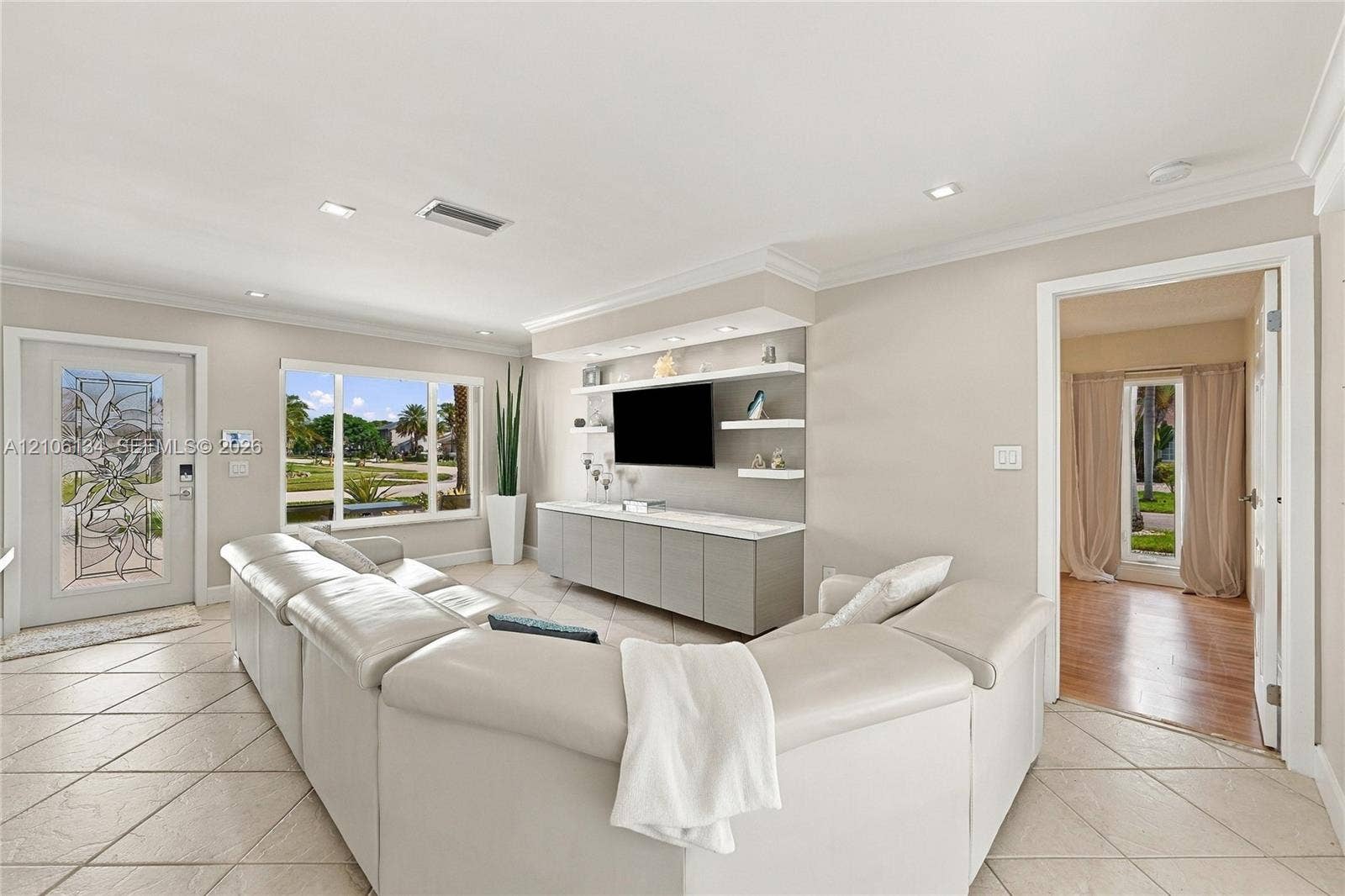 Living area featuring large-format diagonal tile flooring, custom built-in media cabinetry with integrated display shelving, crown molding, and a decorative glass-panel front door
