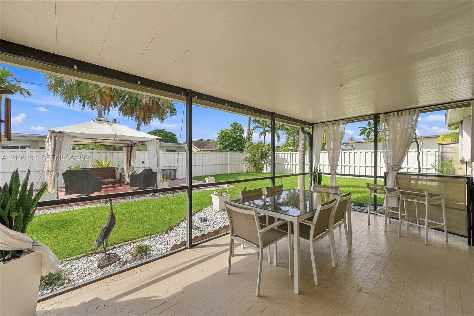 Screened-in patio with tile flooring and a covered ceiling, overlooking a private backyard featuring a manicured lawn, white vinyl fencing, and a detached gazebo