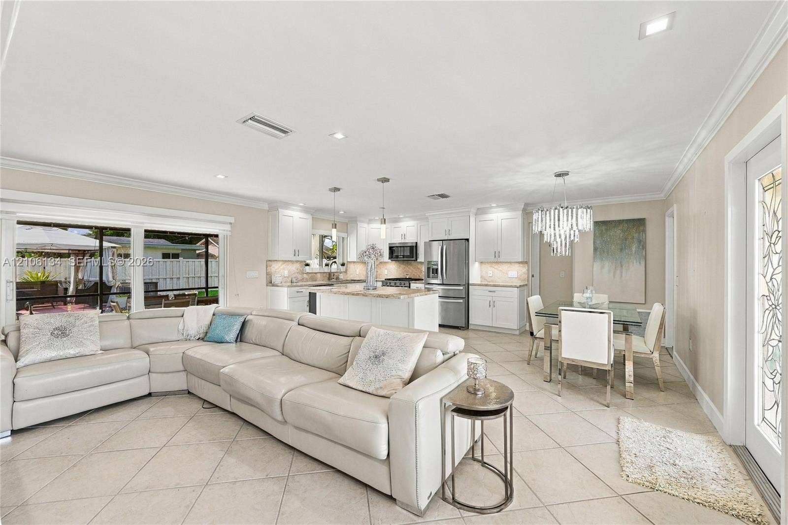 Open-concept interior featuring light-colored tile flooring, white shaker-style cabinetry, stone countertops, stainless steel appliances, and a tiered crystal chandelier